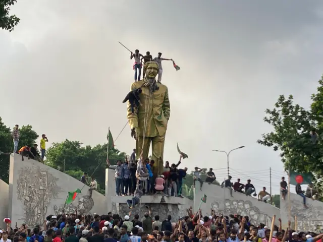 A statue of Sheikh Mujibur Rahman with several protestors standing on top as it is being vandalised