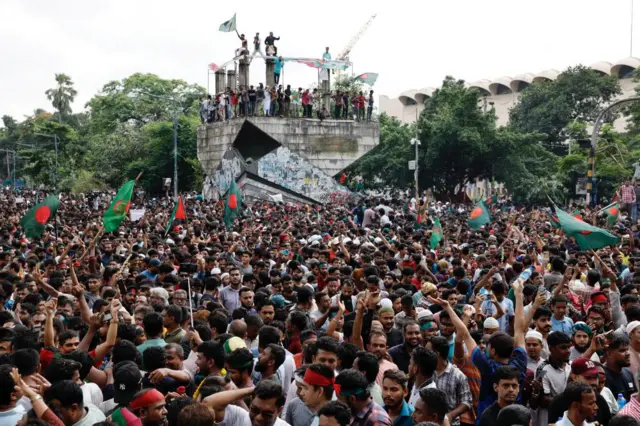 A large crowd in Dhaka celebrating the resignation of the prime minsiter. Lotds of flags are being waved and several are stood on a monument.