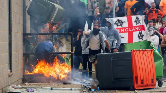 A man throws a wheelie bin as others are set alight as crowds look on