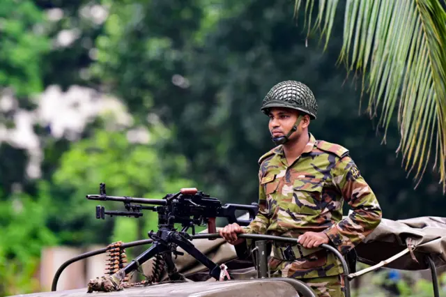 A soldier in camouflage combat uniform stands on the back of a truck next to a mounted machine gun.