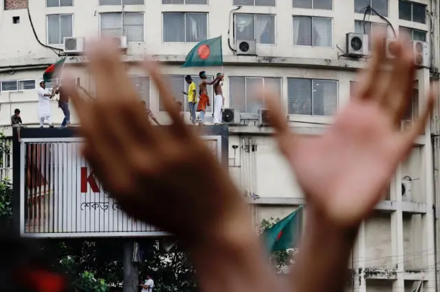 Close up of blurred hands about the clap with a background in focus of protestors standing on a large billboard waving flags of Bangladesh