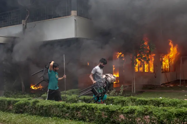 Two men carry chairs in front of a burning building