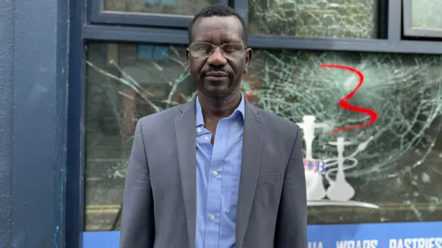 Mohammed Idris stands in front of a smashed glass window