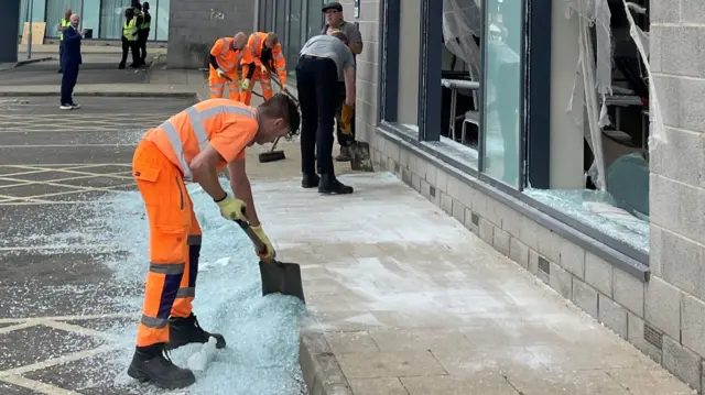 Men dressed in orange use spades to clear up shatter glass