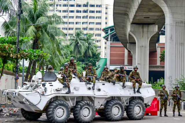 Armed soldiers holding rifles sit on the side of a white armoured personnel carrier.