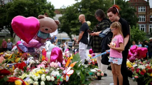 People standing in front of flowers and balloons at vigil in Southport