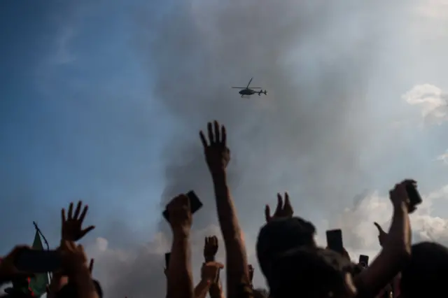 Hands raised in smoke-filled air as a helicopter flies overhead.