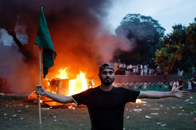 A protester with a Bangladeshi flag stands in front of a burning vehicle at ex-PM Sheikh Hasina's official residence in Dhaka, Bangladesh. Photo: 5 August 2024