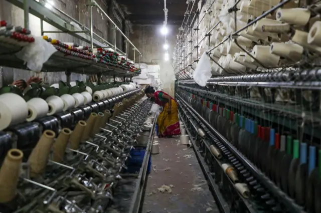 A woman in a colourful dress bends down between rows of yarn and thread in a factory.