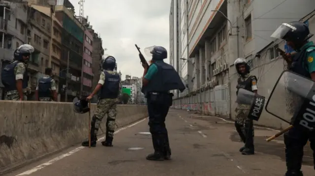 Six police officers in Bangladesh wearing riot gear, the one in the middle is holding a rifle, pointed to the sky