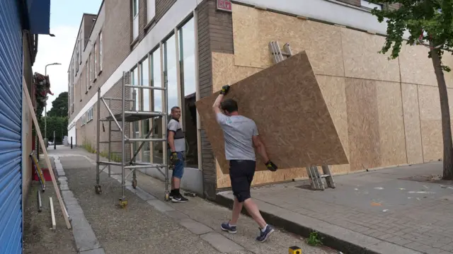 Man walks towards a man stood outside a building's window while holding a board