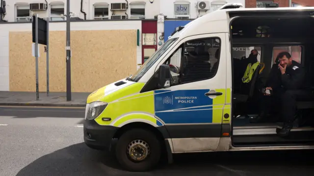 A police van with its back door open is in front of a boarded up shop. A policeman sits in the back of the van