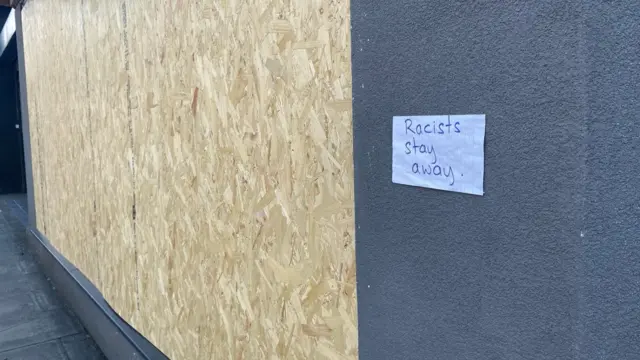 A boarded up window with a piece of paper pinned next to it with Racists stay away written on it in black ink