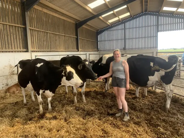 Florence Turley stood in from of a herd of black and white cows wearing shorts and boots