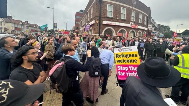 A crowd of people on a Harrow street. One person holds a 'stop the far right, refugees welcome' sign