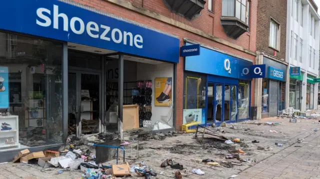 Landscape shot of high street filled with debris. Shoezone store has window smashed and rubbish outside