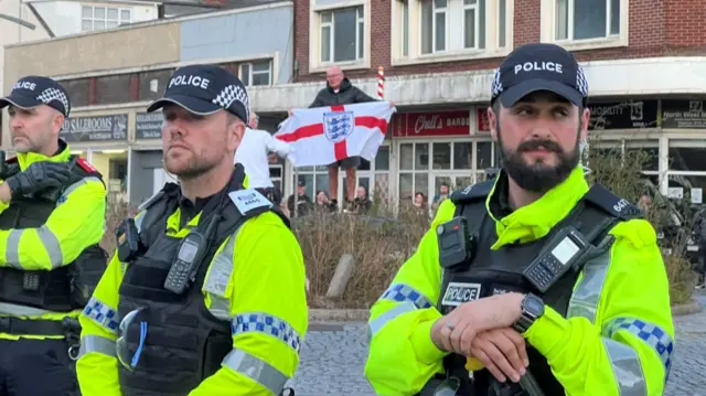 Police stand by as a man behind them holds up an England flag