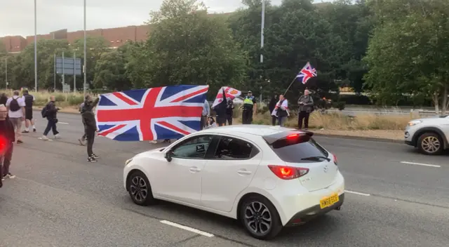 Protesters holding a large Union Jack flag block a car on a main road