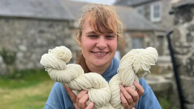 Elsa poses with her spun wool in front of stone farm buildings