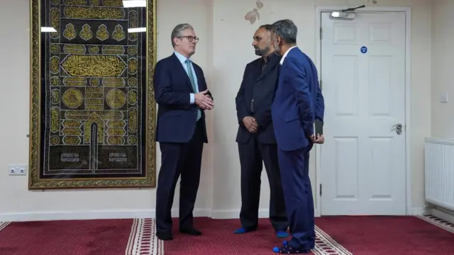 Keir Starmer stands in a mosque and speaks with two representatives with their shoes off