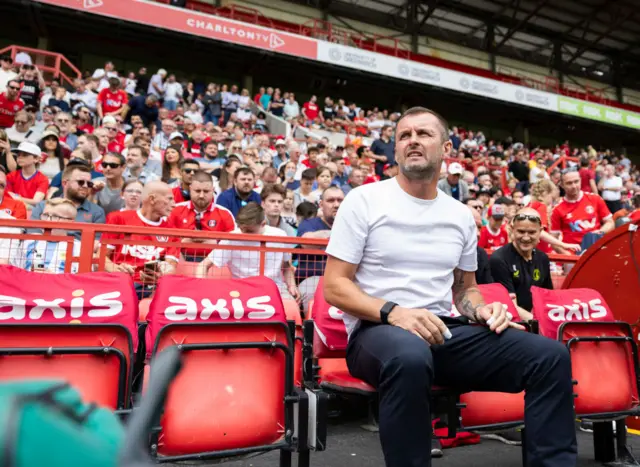 Charlton boss Nathan Jones sits in the dugout with fans behind him