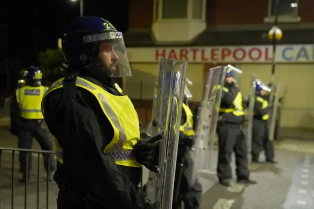 A line of police officers in riot gear standing in a street with a shop sign in the background with the word Hartlepool