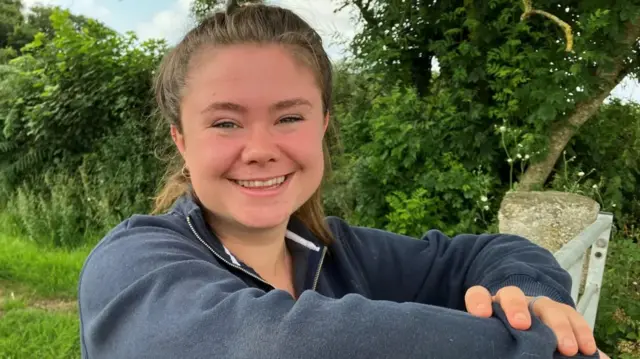 A smiling Maria Warne-Elston standing at a gate wearing a blue jumper