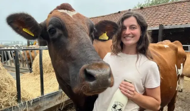 A smiling Erin Shreeve standing with a cow and holding a bottle of milk
