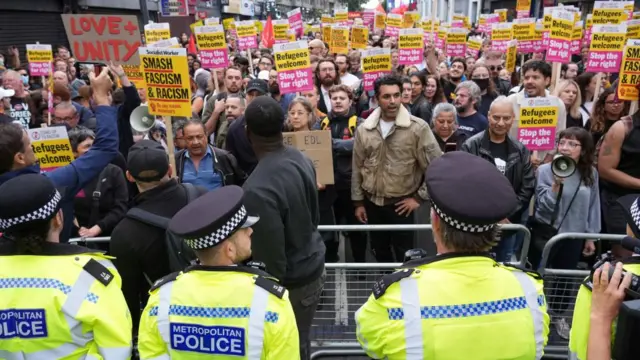 Police officers stand outside a peaceful protest in Walthamstow while people hold signs saying 'Refugees Welcome'