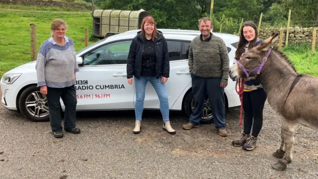 Katie, her parents, and the BBC's Steph Finnon stand in front of a Radio Cumbria-branded car with a donkey