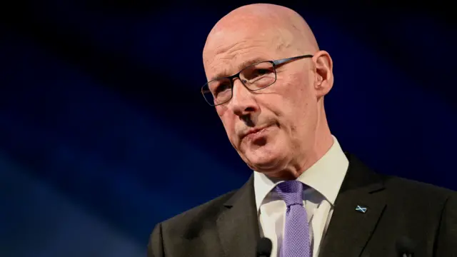 John Swinney looks off to the side of the camera. He is wearing black glasses and a suit with a violet tie