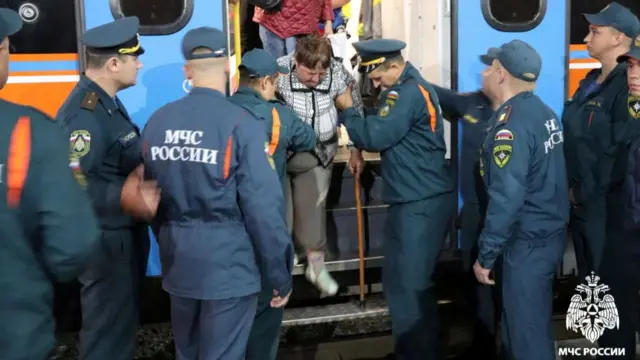 Russian Ministry of Emergency Situations staff help a woman walking with the aid of a stick off a vehicle