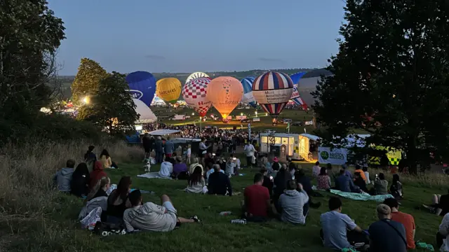 Crowd of people sat watching the night glow. Hot air balloons can be seen in the distance, inflated on the ground, lit up with fire.