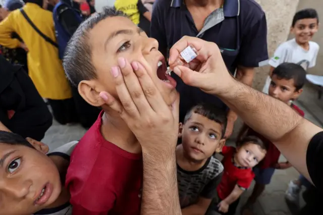 A child looks up as a health worker holds his face and puts drops in his mouth while children are gathered around him