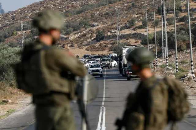 Two soldiers, out of focus in the foreground, looking at a group of cars on the road near where three Israeli police were shot dead