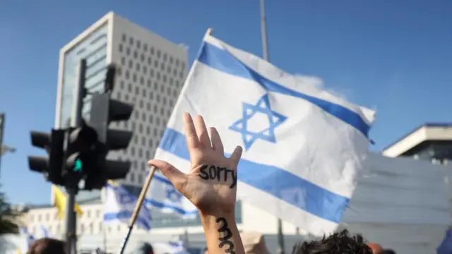 A hand with the word 'sorry' is held up against the backdrop of a demonstration in Jerusalem, while an Israeli flag waves in the background