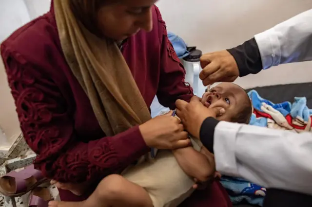 Health worker gives a baby drops into their mouth on hospital bed, with a woman holding the baby