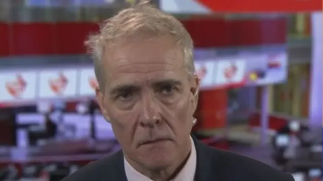 Charlie Taylor headshot, he sits in front of the BBC Newsroom in a suit