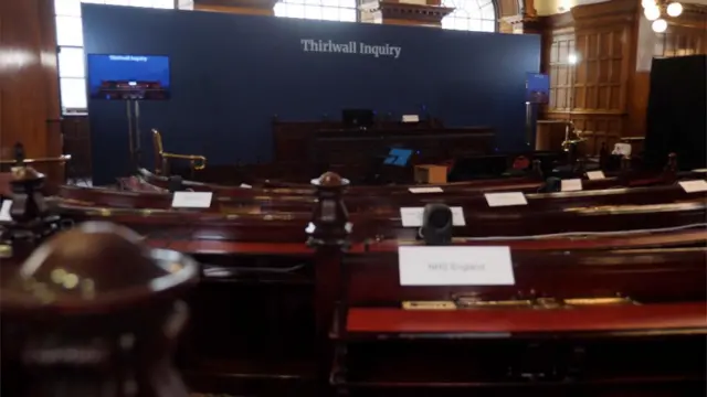 The inside of a wood-panelled inquiry room with a wide dark blue screen with white letters that read 'Thirwall Inquiry'