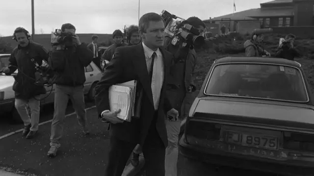 Pat Finucane carrying a bundle of papers being followed by cameramen, the image is in black and white