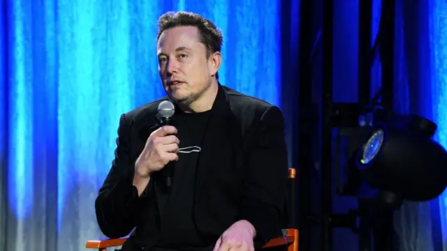 Elon Musk sitting holding a microphone