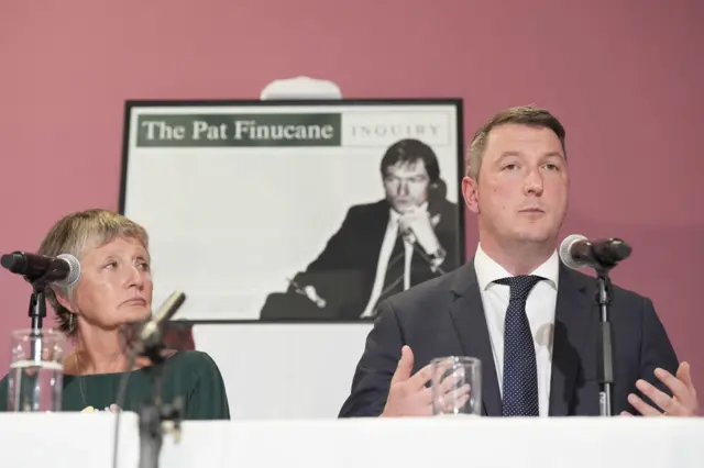 Geraldine in a green dress looking at her son John who is wearing a blue suit and speaking into a microphone