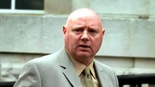 William Stobie, a bald man wearing a grey suit, yellow shirt and yellow tie looks towards the camera