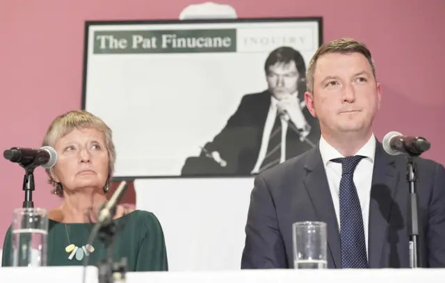 Geraldine wearing a green dress sitting next to John a suit looking out to press