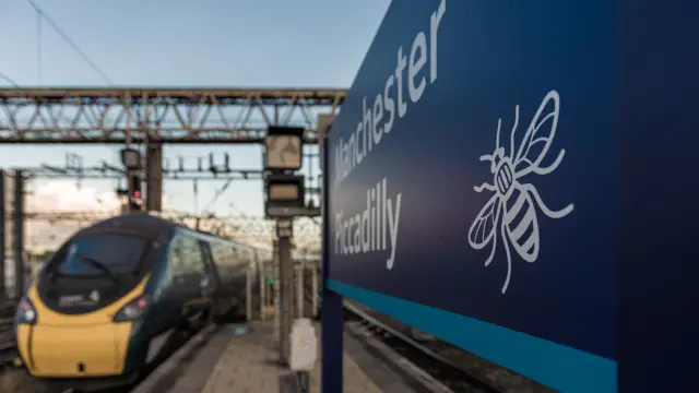 The Manchester bee on a sign that says Manchester Piccadilly. A train moves in the distance