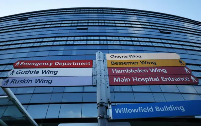 A photo taken from below of a signpost showing directions to eight NHS wards at a hospital. A large, glass panelled building stands behind.