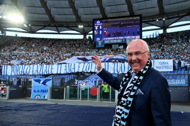 Sven Goran Eriksson look into the camera while waving at a stand full of people on a visit to former club Lazio