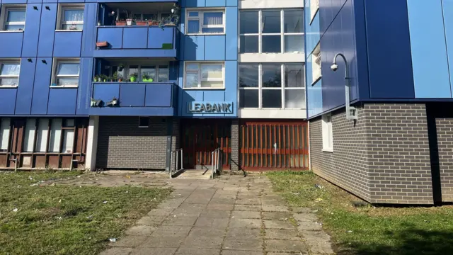 The entrance of Leabank, a pavement leads towards a wooden brown entrance. The building, covered in in blue cladding, is seen above it. The words "LEABANK" are written above the door.