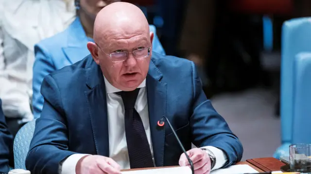 Vassily Nebenzia sits at a table speaking into a microphone. He is wearing a navy suit and black tie