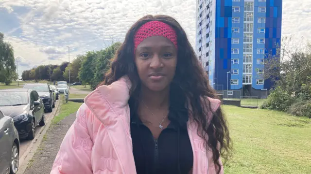 A woman wearing a pink coat and a necklace that with the letter M is standing in front of Leabank